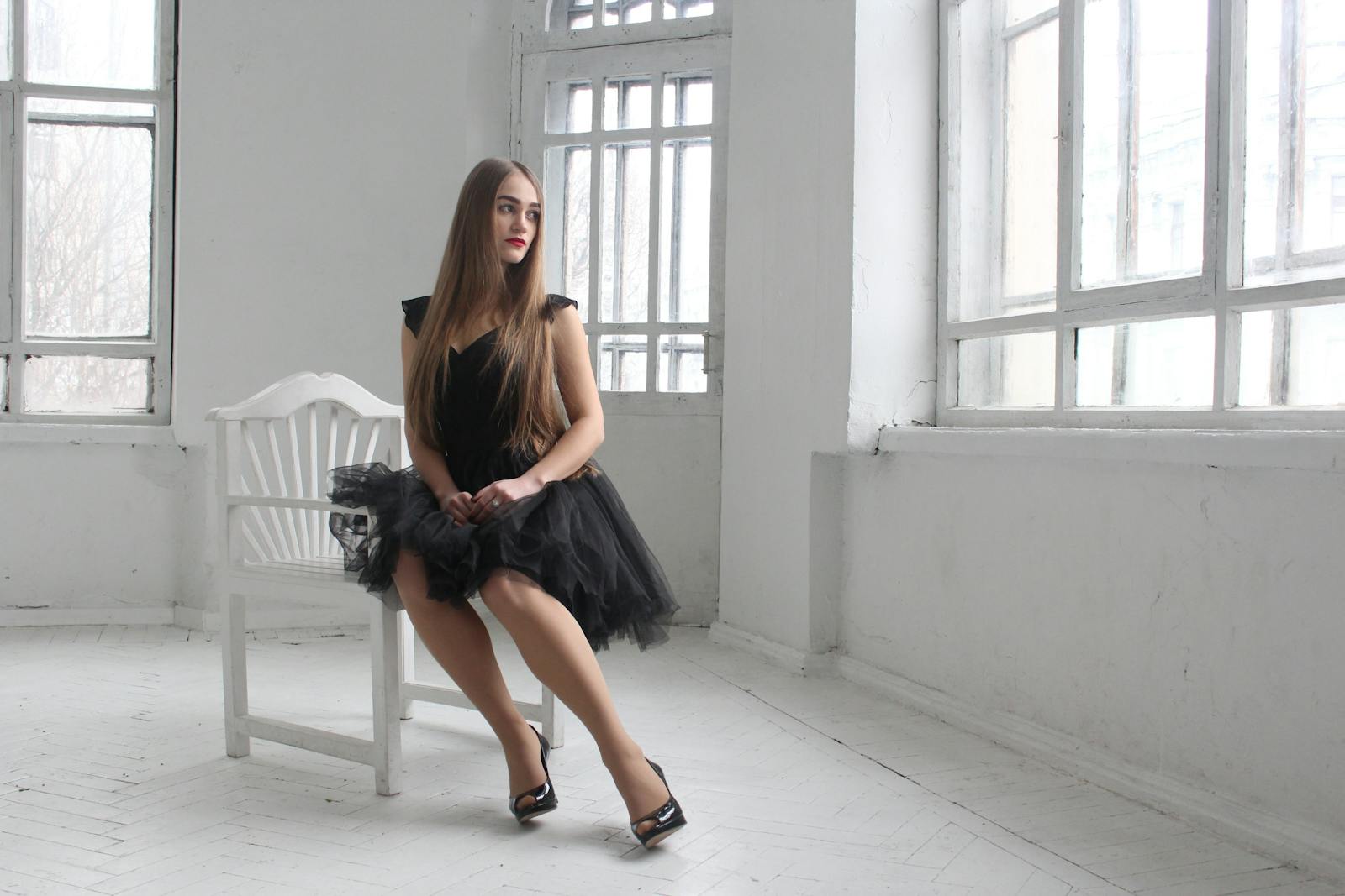 A young woman in an elegant black dress poses in a bright room with large windows, showcasing beauty and style.